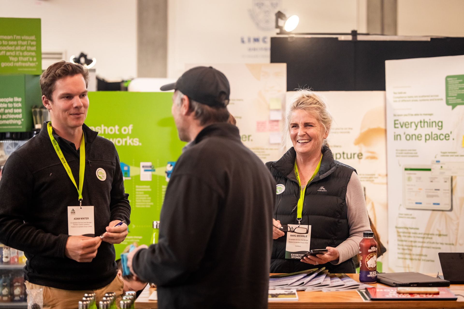 Three people converse at a booth during an event. The woman smiles and holds a phone. The booth has promotional posters.