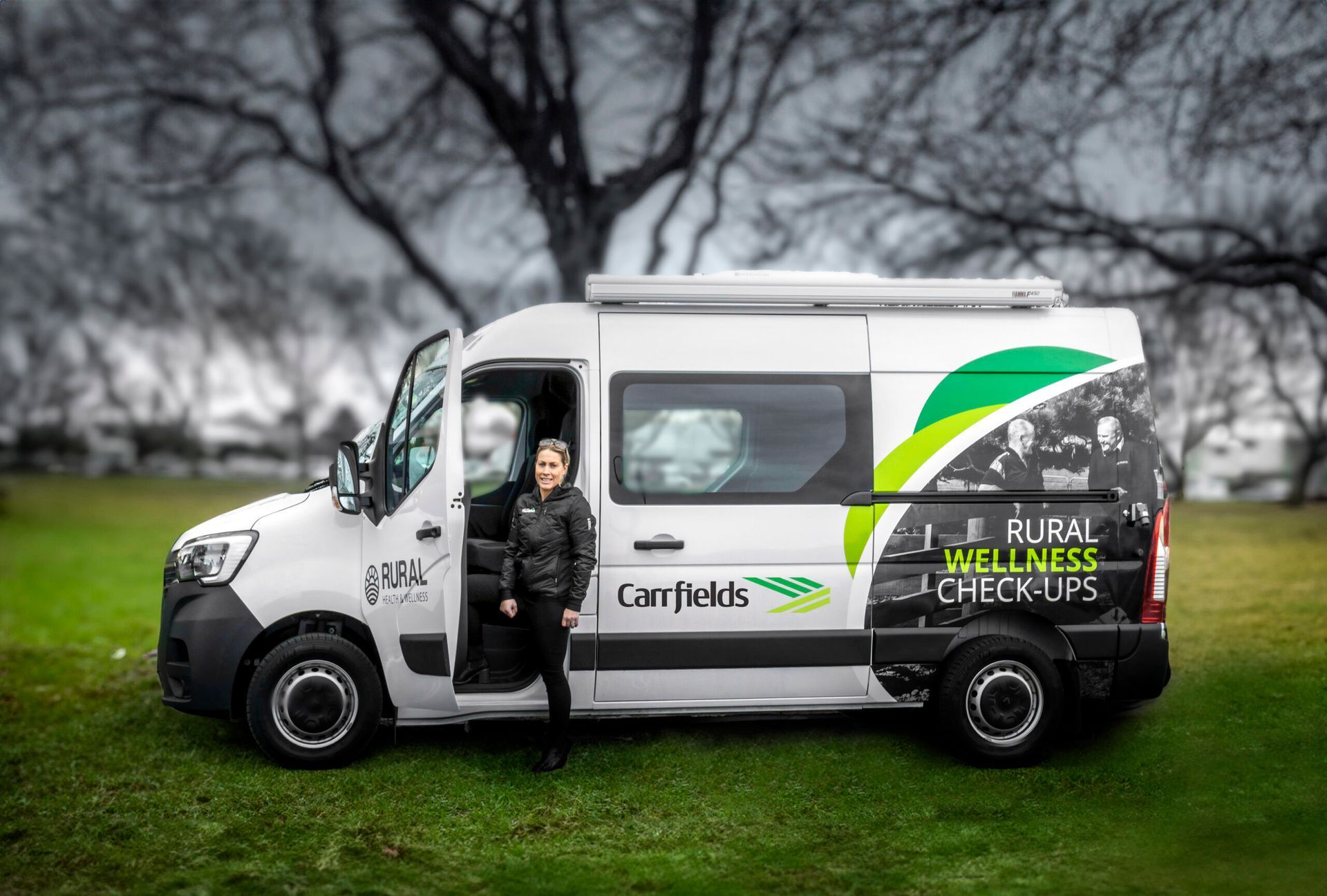 Woman stands in front of a white camper van with green and black logo on grass.