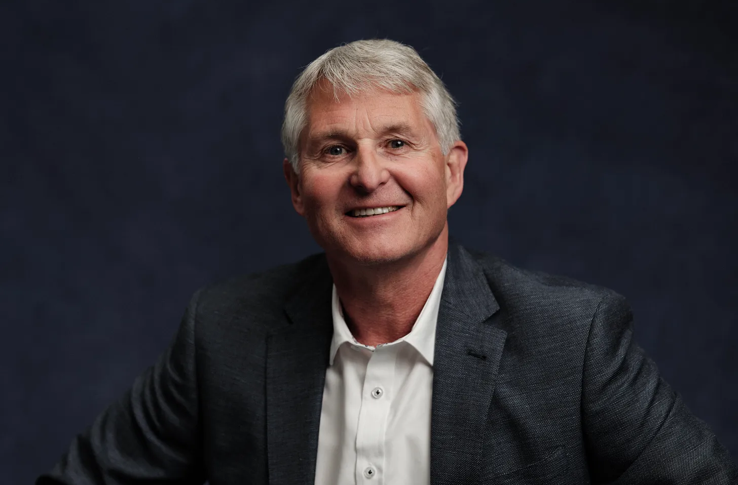 Smiling man in a dark suit jacket and white shirt against a navy blue backdrop.
