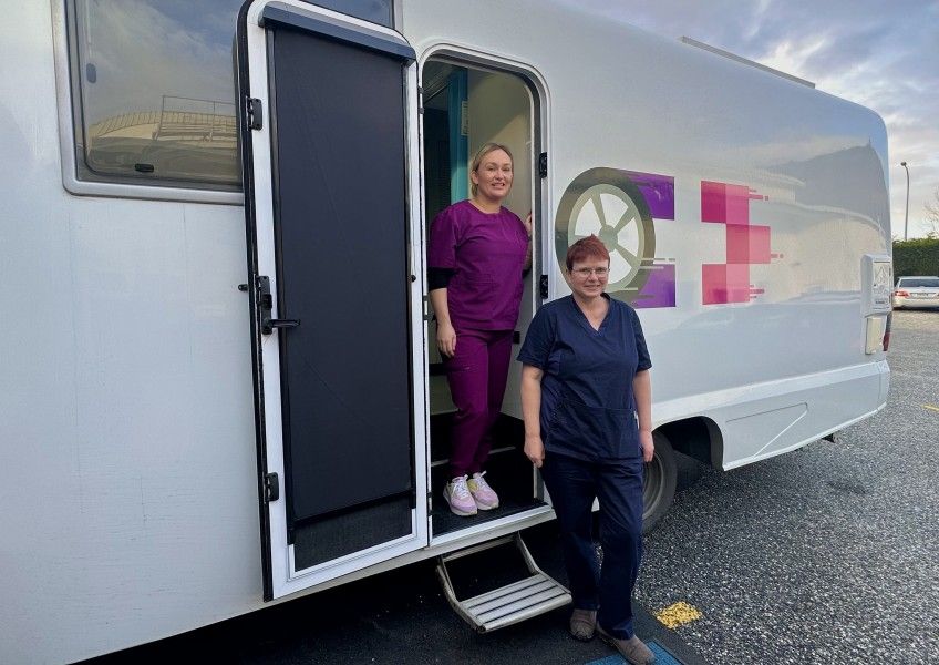 Two medical professionals in scrubs stand outside a mobile clinic. One is exiting, the other is by the door.