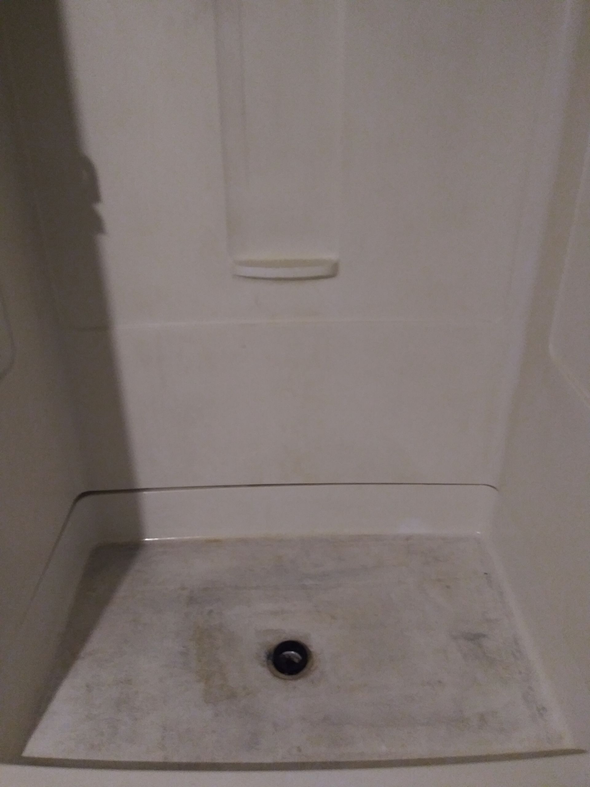 White shower stall, soap dish, dark drain, stains on the floor.