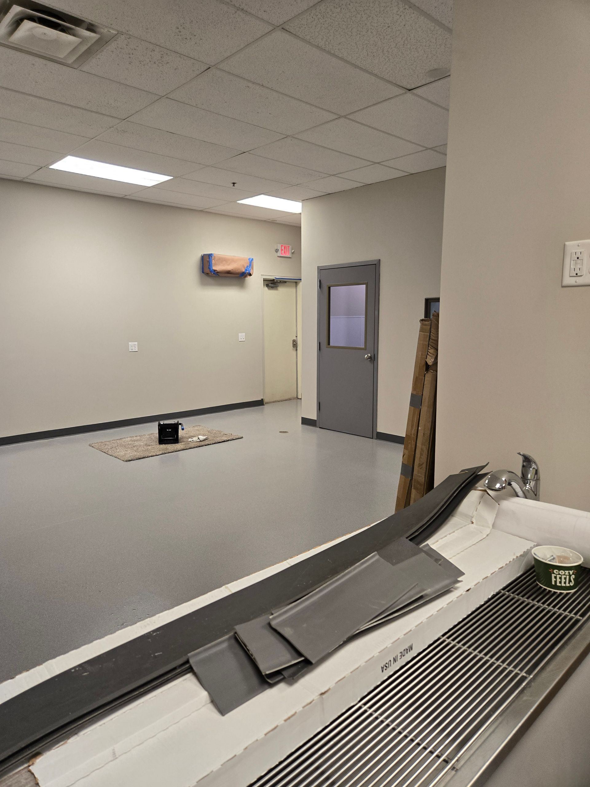 An empty, gray-floored room with a doorway and a sink in the foreground. Construction materials sit on the sink.