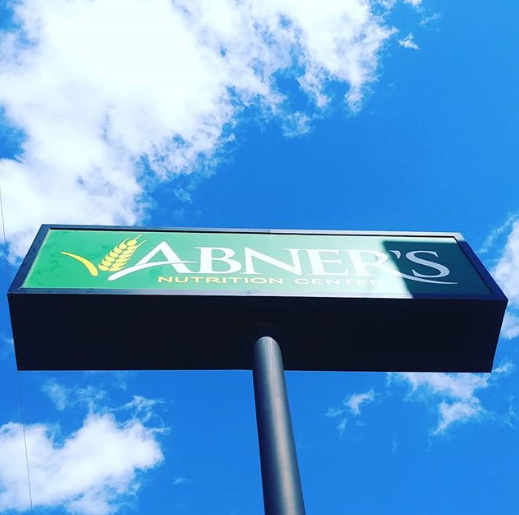 Sign for Abner's Nutrition Shop, green and white on black frame, against a blue sky with clouds.
