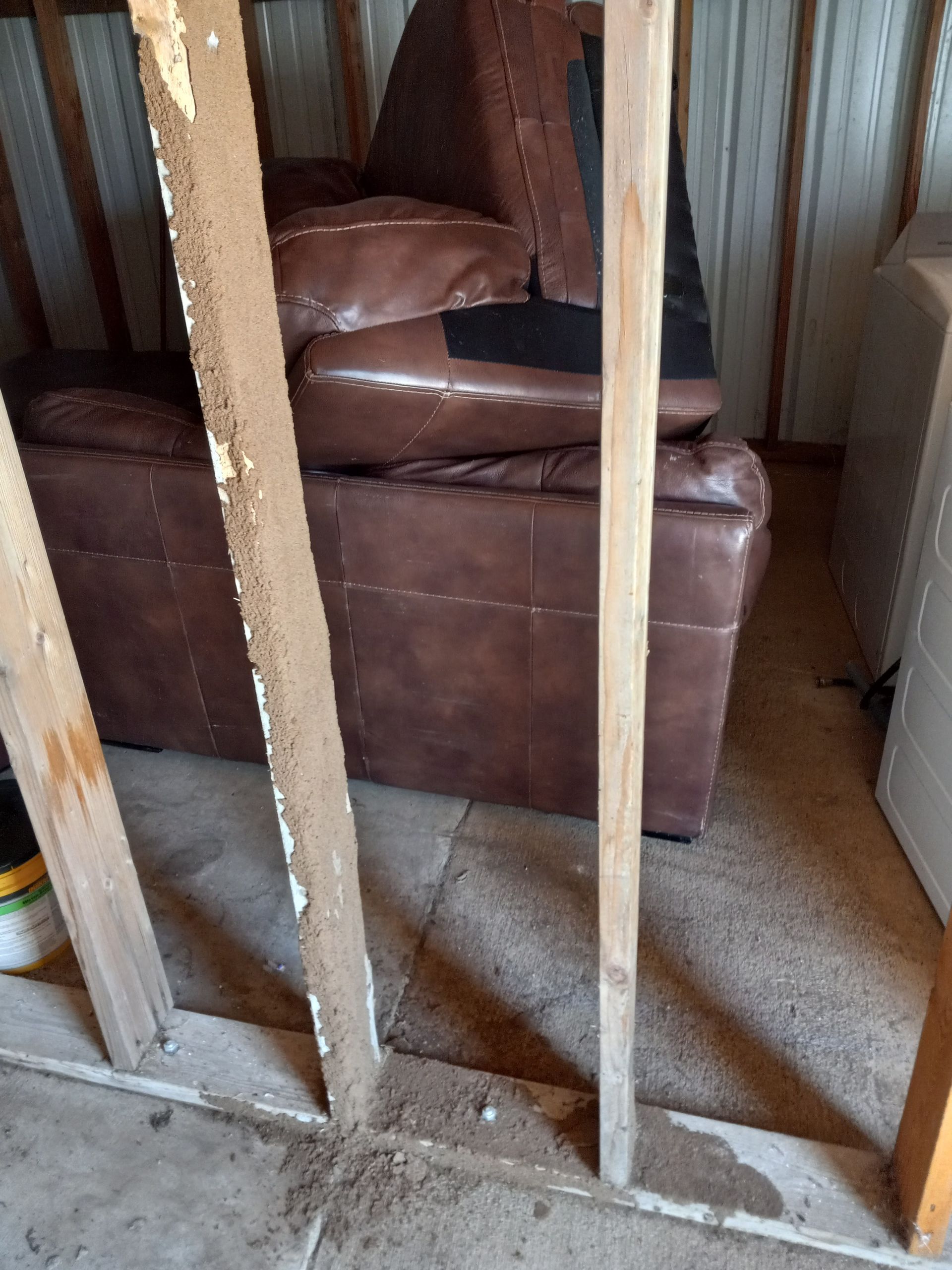 Brown leather couch inside a shed. Wooden studs with debris on the floor in front.