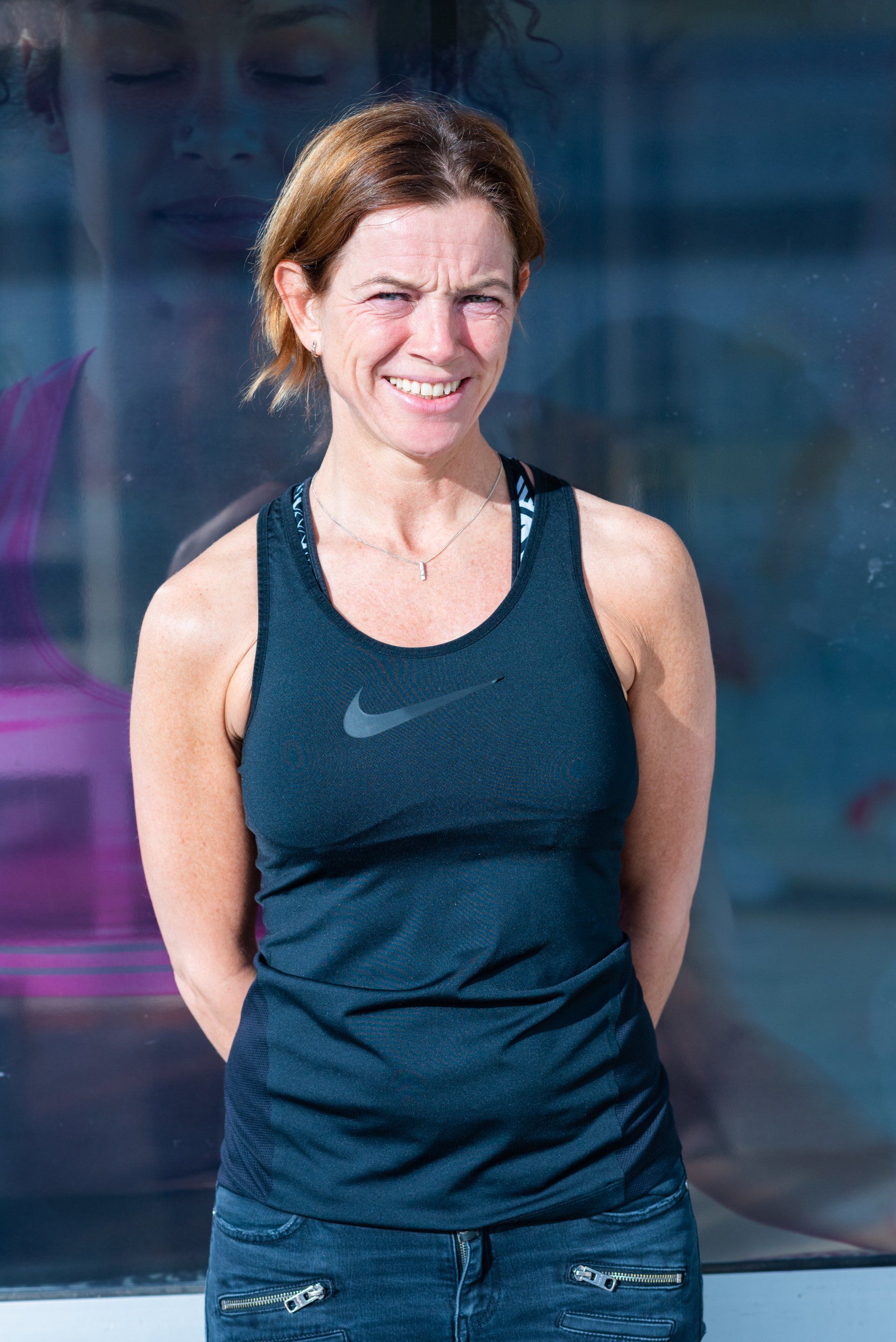 Een vrouw, gekleed in een zwarte nike tanktop en spijkerbroek, glimlacht.