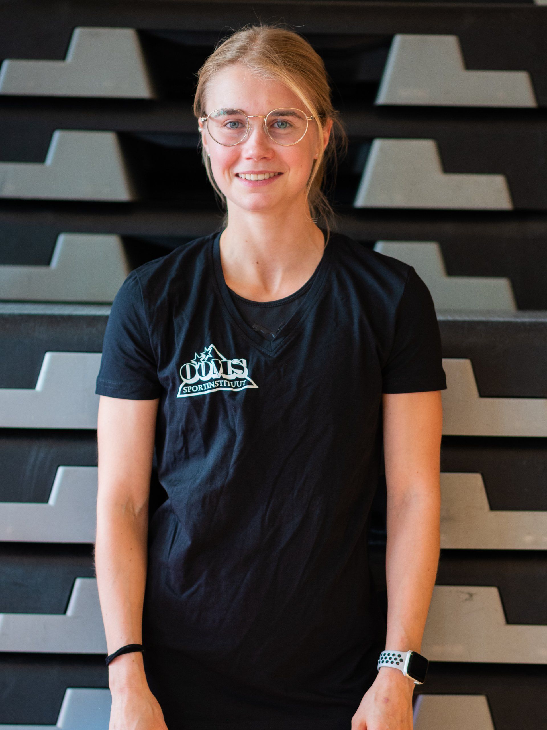 Een vrouw met een bril en een zwart shirt staat voor een muur.