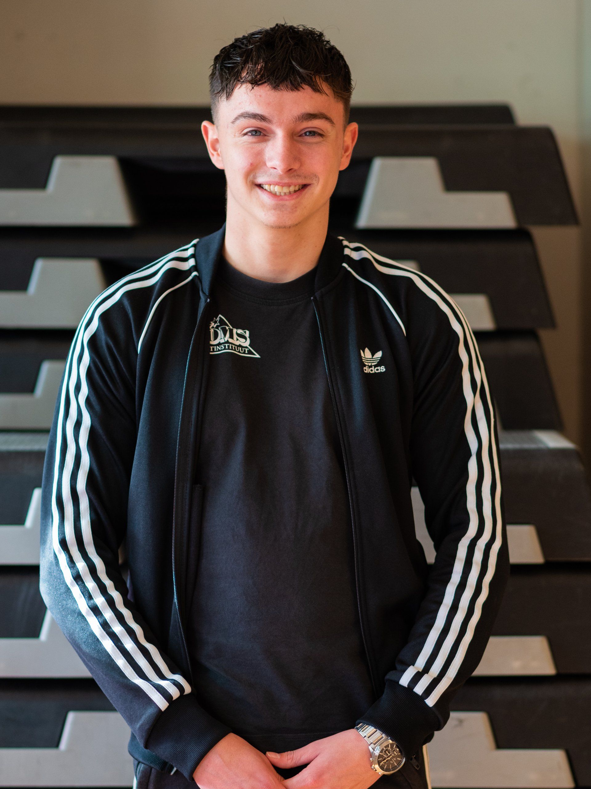 Een jonge man, gekleed in een zwart adidas-jack, glimlacht naar de camera