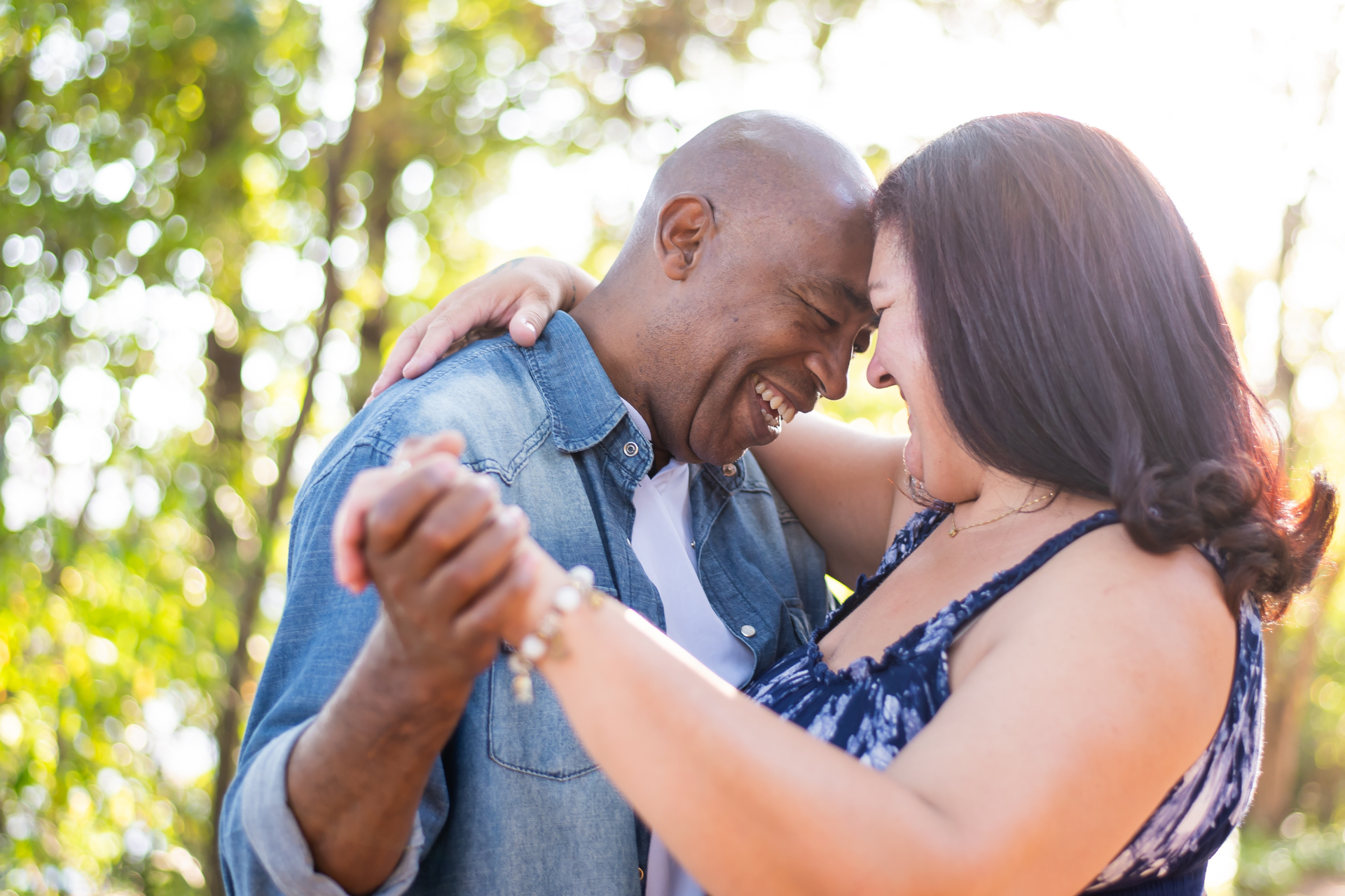 Couple dancing outdoors, smiling, embracing, against a blurred green backdrop of trees.