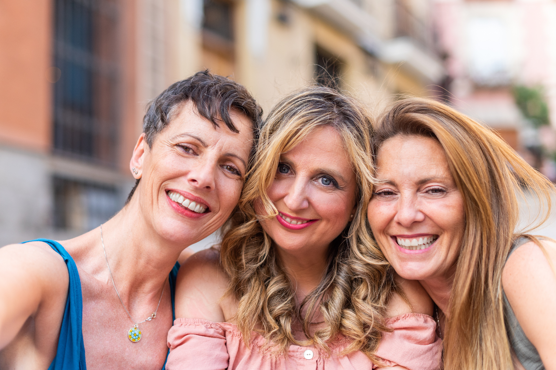 Three smiling women pose for a selfie in a city setting.