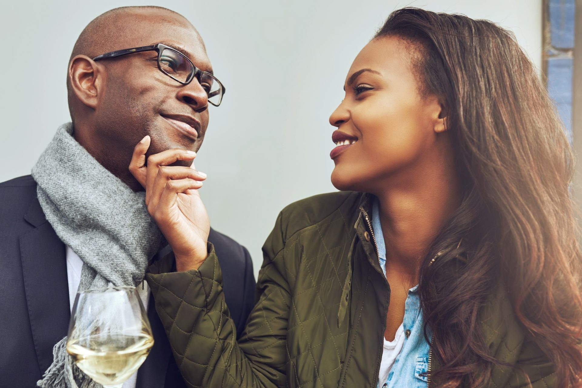 Man holding a glass of white wine gazes at a woman, she touches his chin, both smiling. Outdoors.