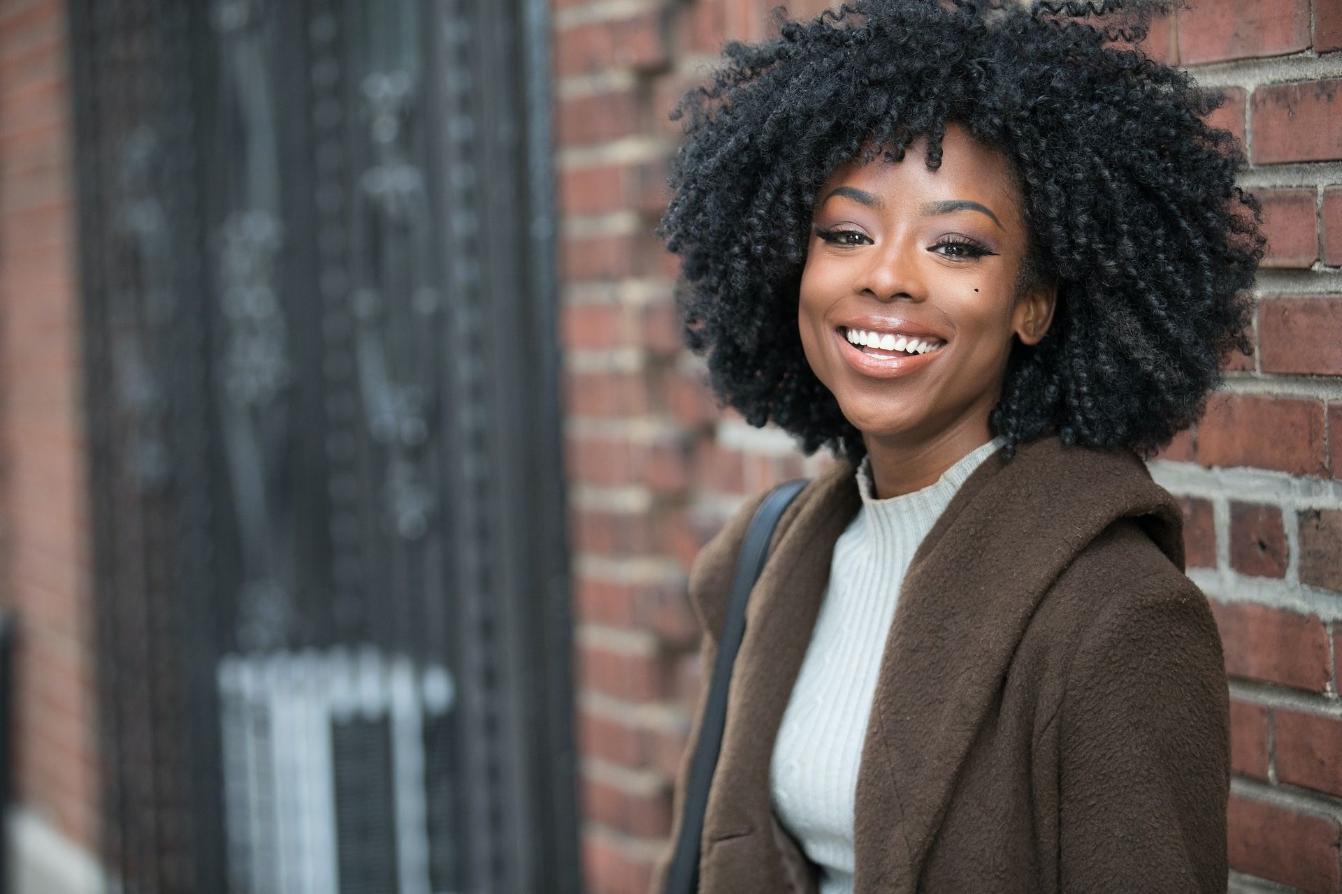Woman with large, curly hair smiles, leaning against a brick wall. She wears a brown coat and light sweater.