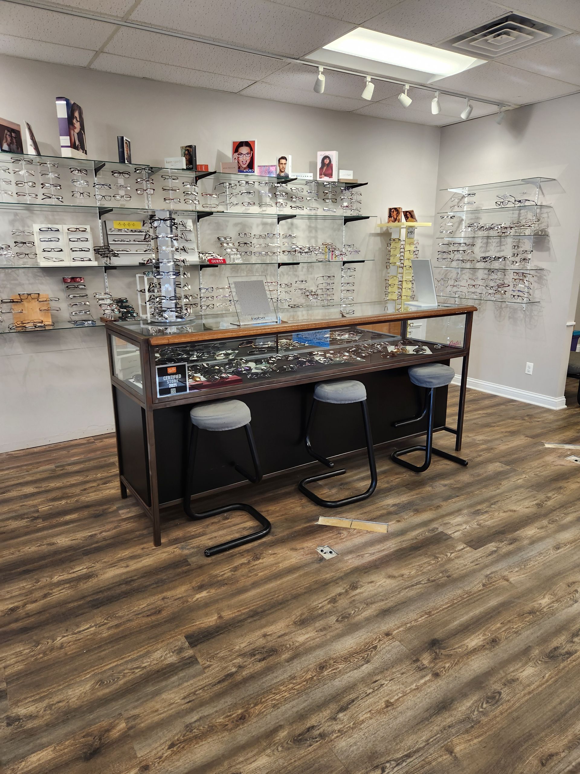 An optometrist's office with eyeglasses on display. Dark wooden flooring, a countertop with stools, and overhead lighting.