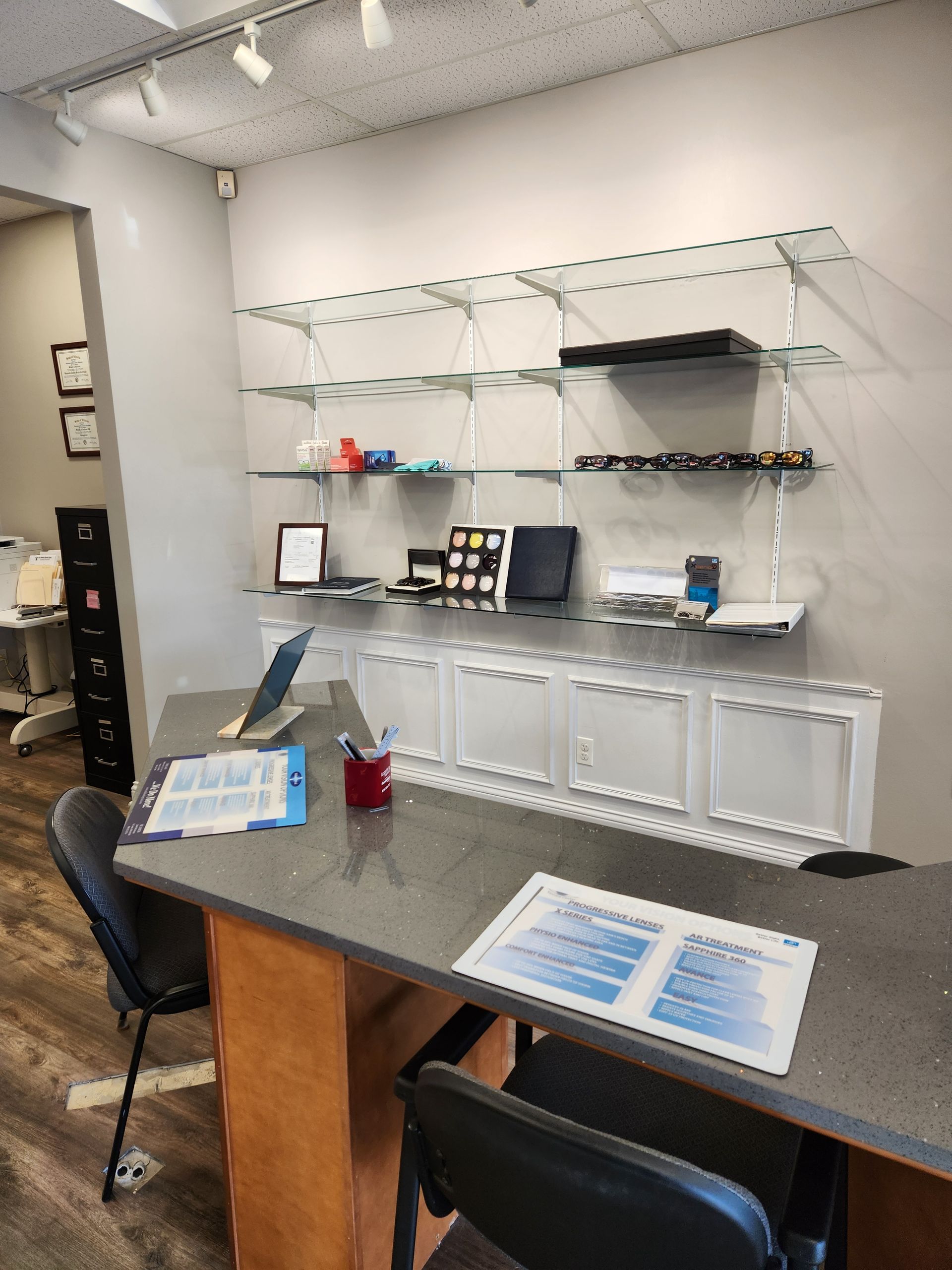Reception desk in an office with gray countertop, glass shelves with eyewear, and paperwork.