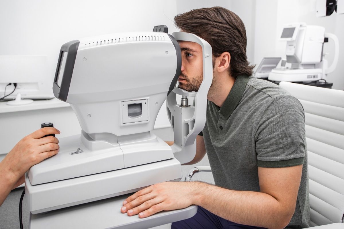 Man using eye exam machine in a medical office, with another person's hands operating it.