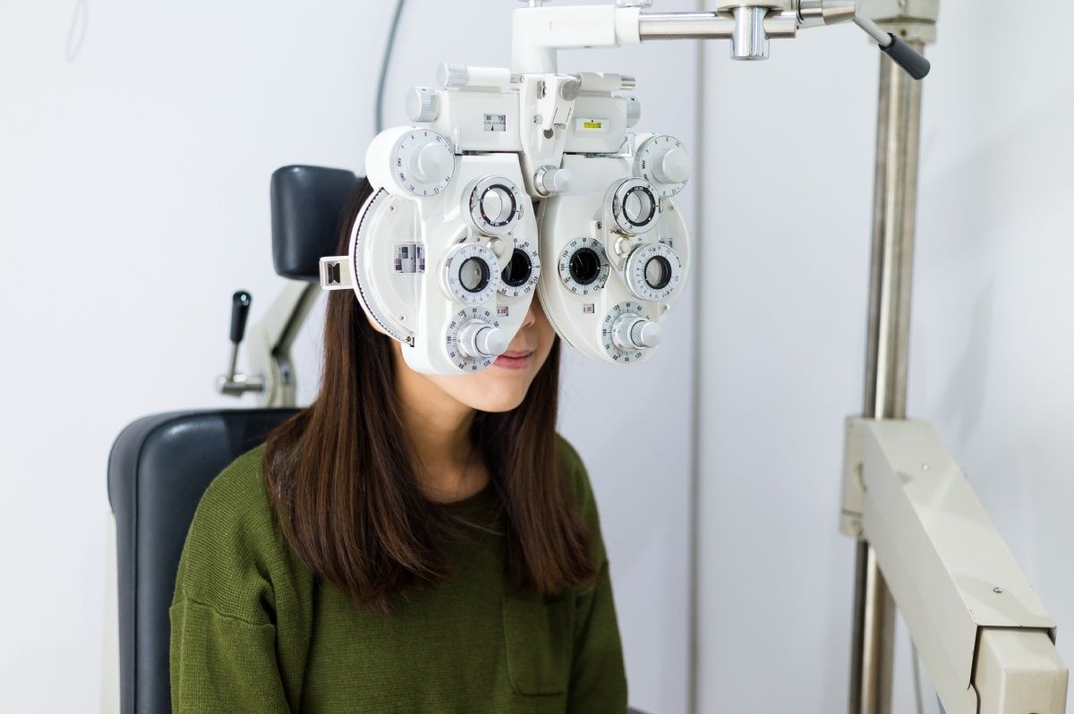 Woman undergoing an eye exam, seated with her face in a phoropter in a white examination room.