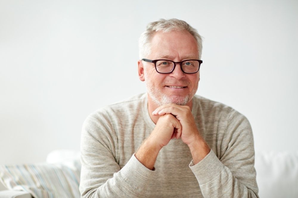 Man with gray hair and glasses smiles, rests chin on hands, neutral-colored sweater, white background.
