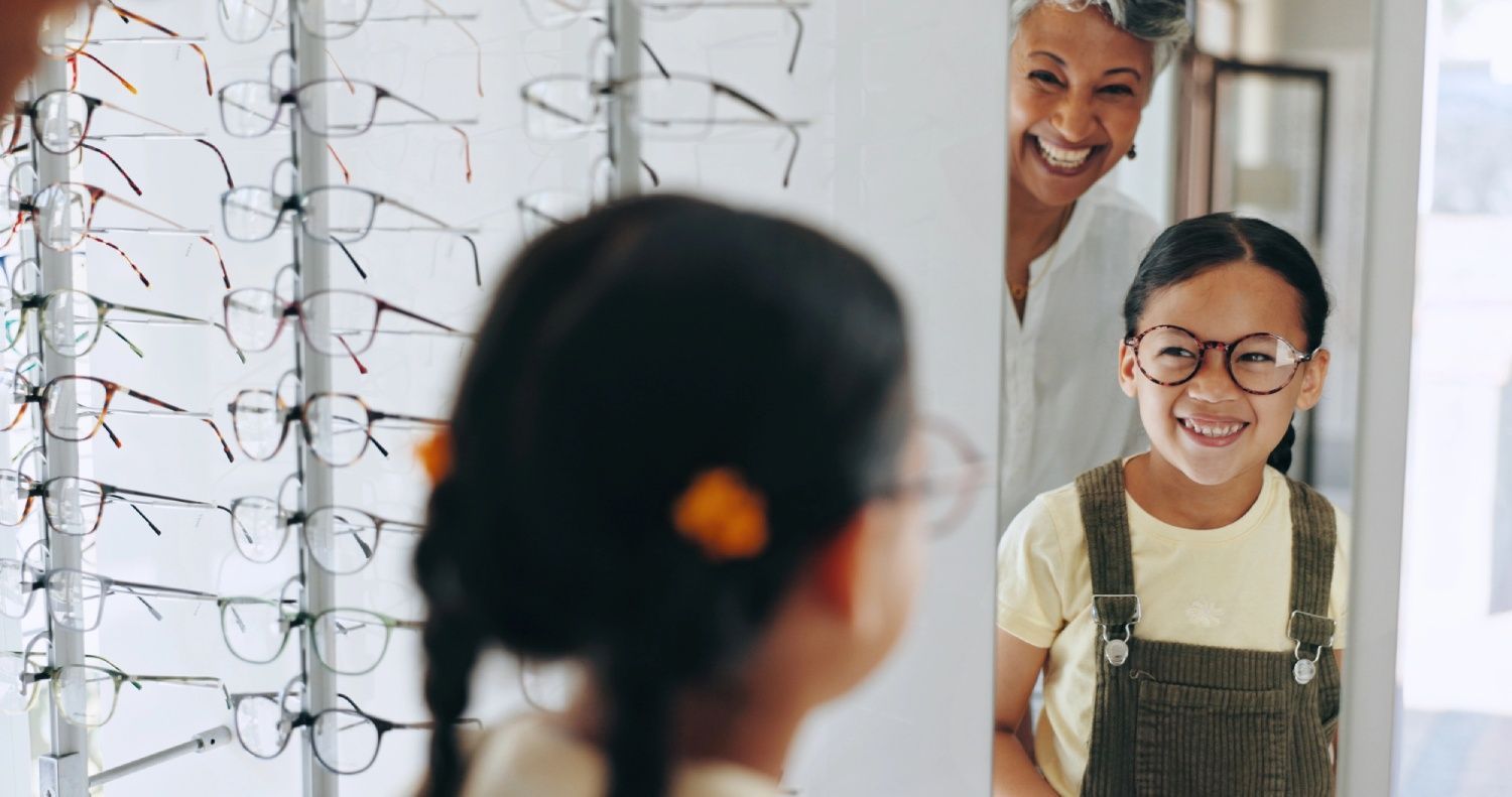 Girl in new glasses smiles at mirror with a woman standing beside her in an optical shop.