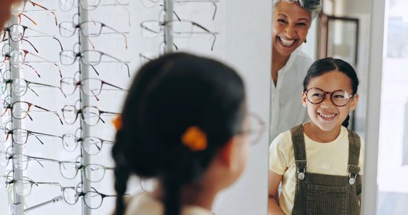 Girl in new glasses smiles at mirror with a woman standing beside her in an optical shop.
