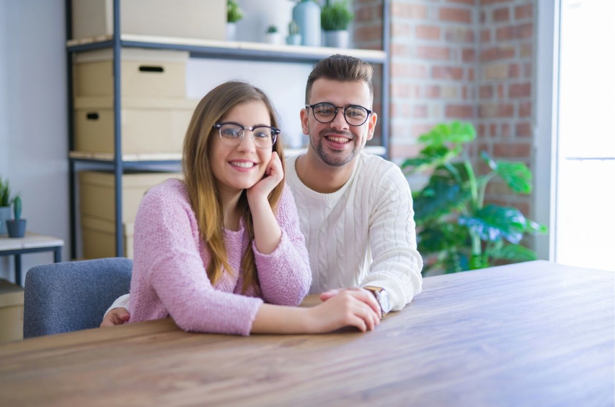 Couple with eyeglasses smiling together at a wooden table. The woman rests her head on her hand.