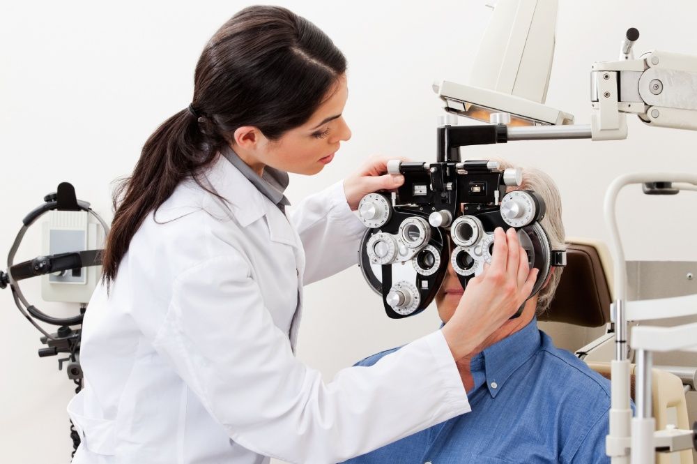 Optometrist examining a patient's eyes with a phoropter in an exam room.
