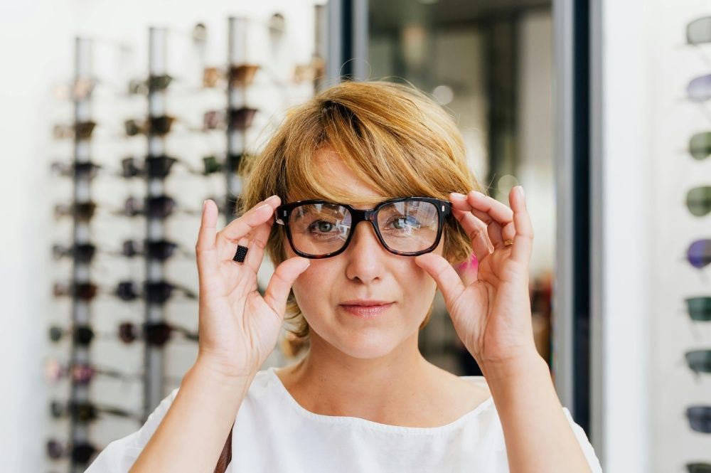 Woman holding up eyeglasses in an optician's shop. Eyeglasses on display in background.