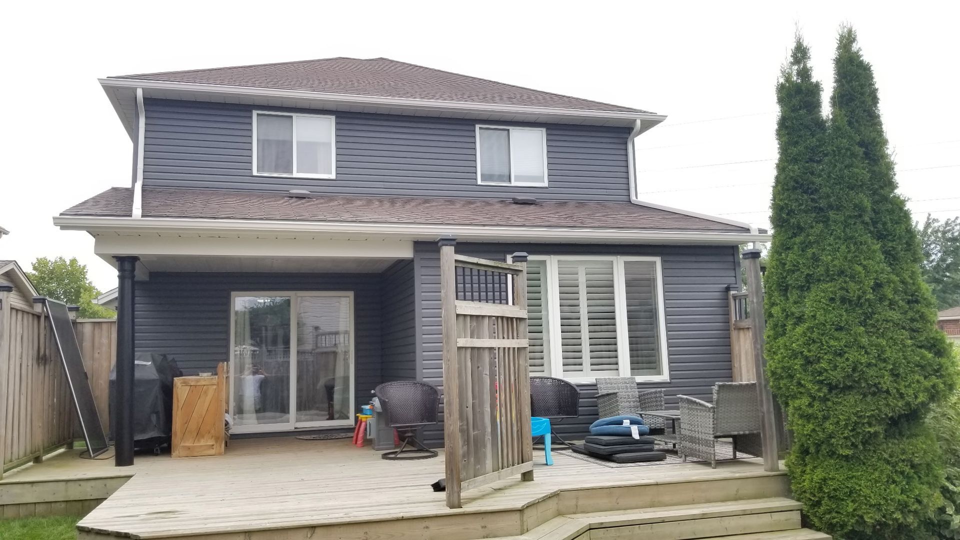 Dark gray house exterior with patio and gate. Tall green tree right. Cloudy day.
