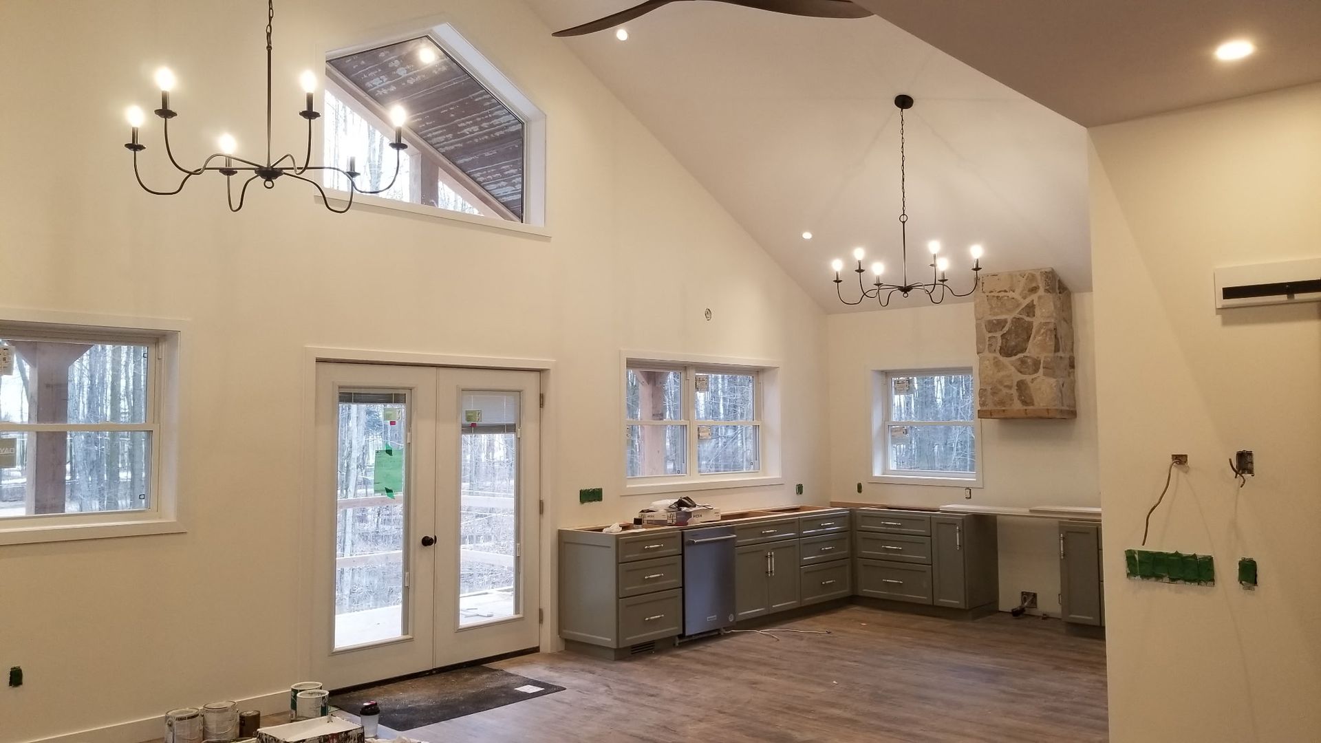 Interior of a house under renovation with tall ceilings, windows, two chandeliers, and gray cabinets.