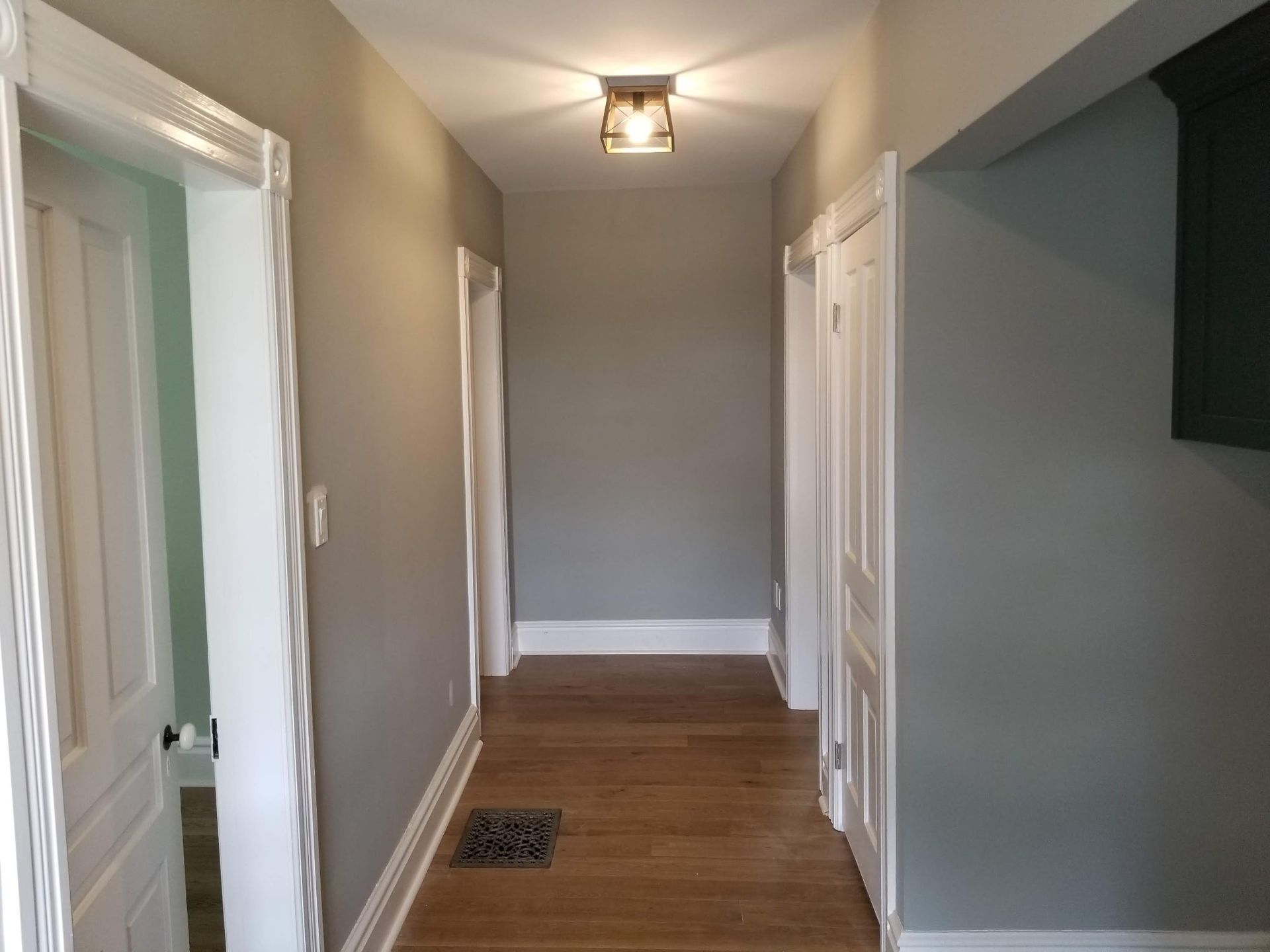 Narrow hallway with gray walls, white trim, wood floor, and doors. A light fixture is mounted on the ceiling.
