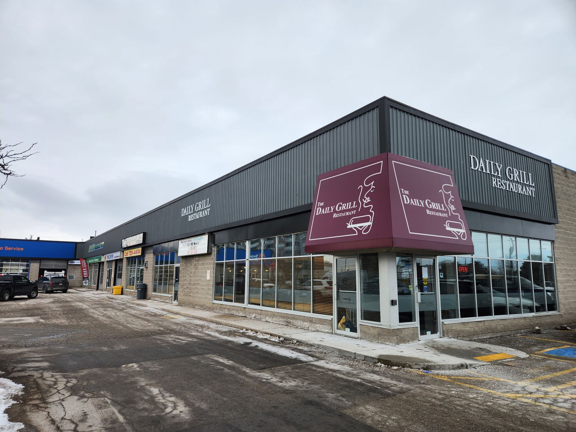 A strip mall with various storefronts, some with signs. The sky is overcast and the ground is covered in patches of snow.