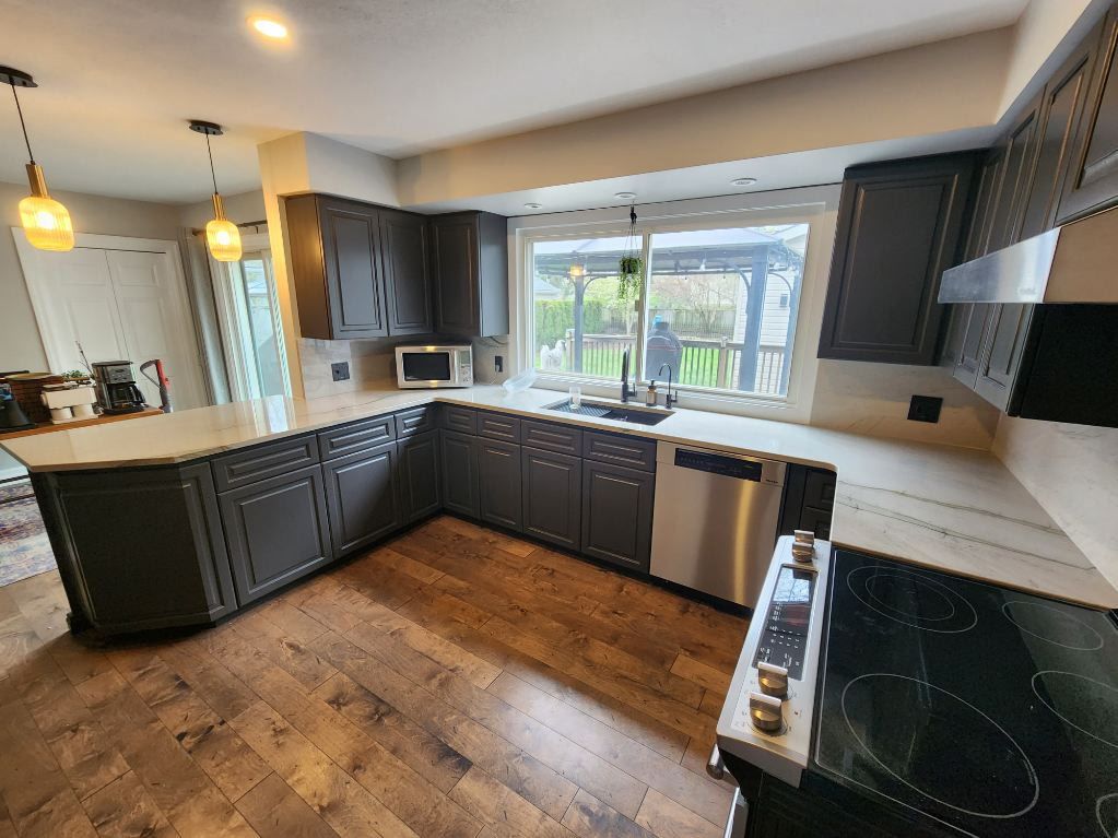 A kitchen with dark gray cabinets, a wooden floor, and a large window overlooking a yard.