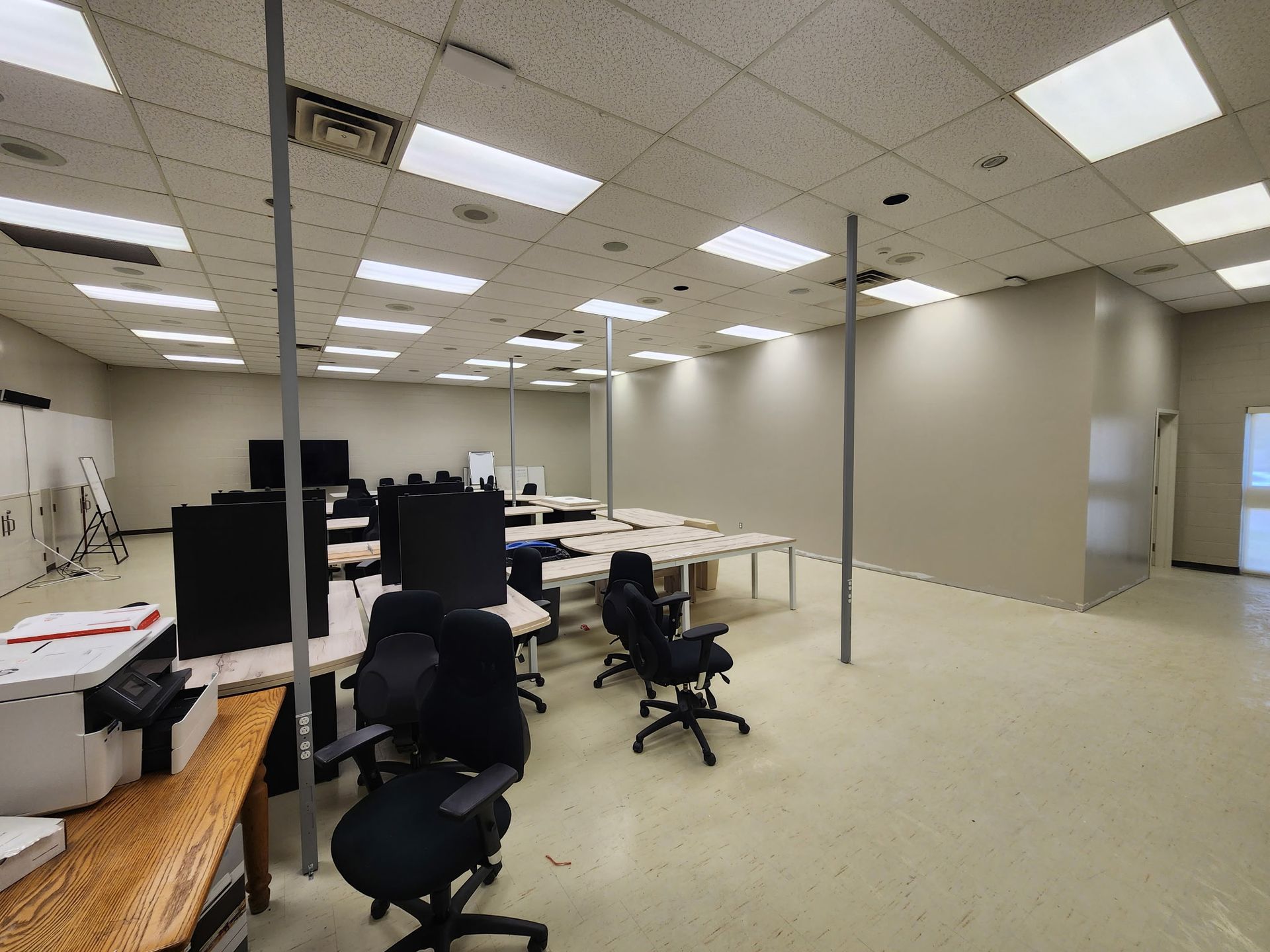 An empty office space with desks, chairs, and support poles. The walls are light beige and the floor is a light tan.