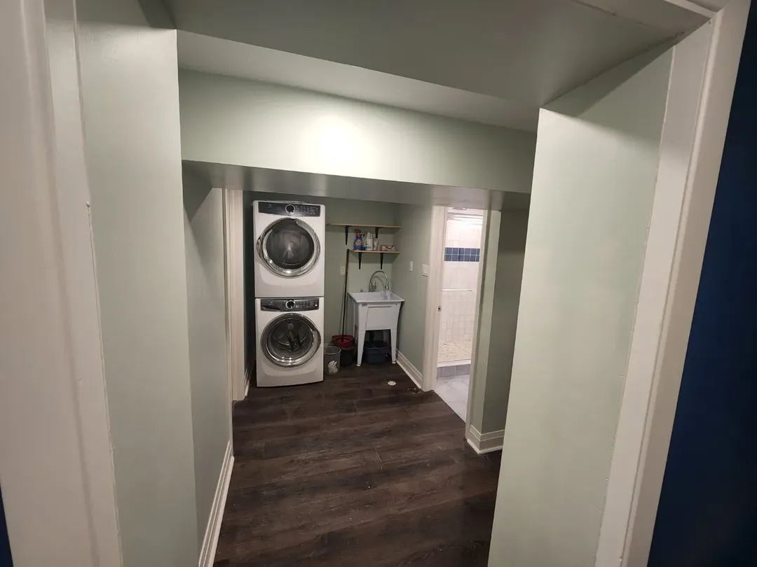 Laundry room with stacked washer and dryer, sink, and doorway. Wooden floor and light green walls.