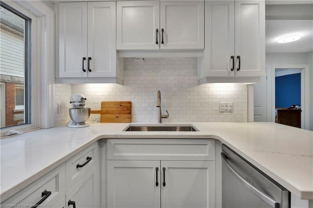 White kitchen with white cabinets, countertops, and backsplash, stainless steel sink and appliances.