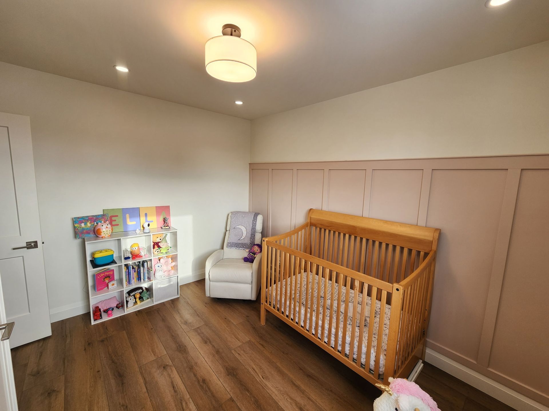 Empty nursery with a wooden crib, white chair, and book shelf against light pink and white walls.