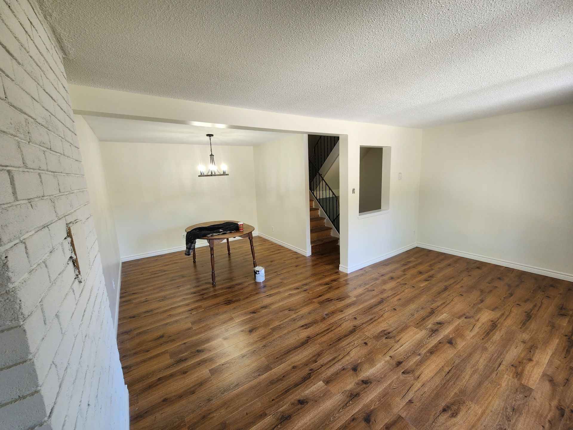 Empty room with wood floors, brick wall, and stairs in the background. Ceiling is textured and walls are light.