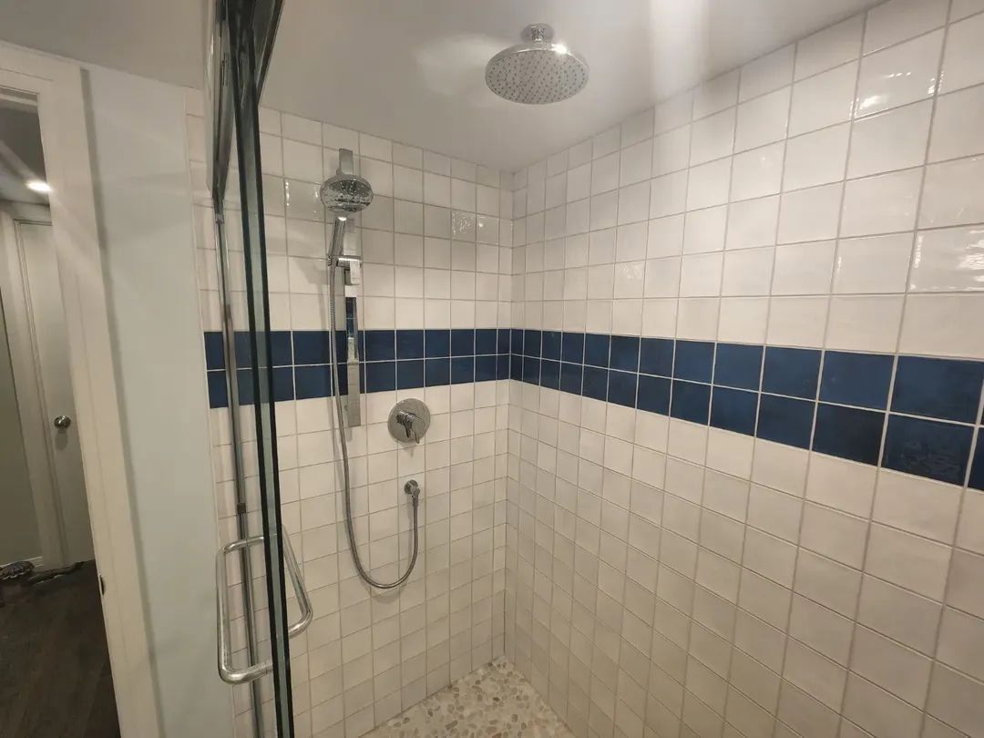 Shower with white tiles, a blue stripe, and silver fixtures. A glass door is on the left.