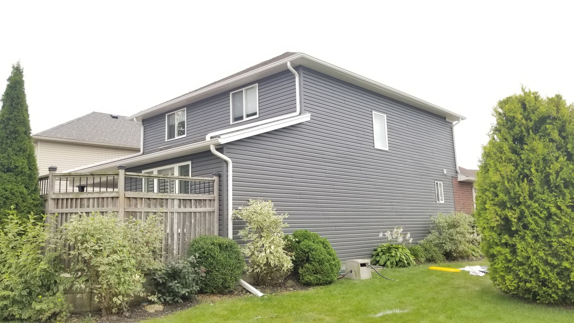 Gray-sided house with white trim, green lawn, wooden fence, and surrounding greenery under a cloudy sky.