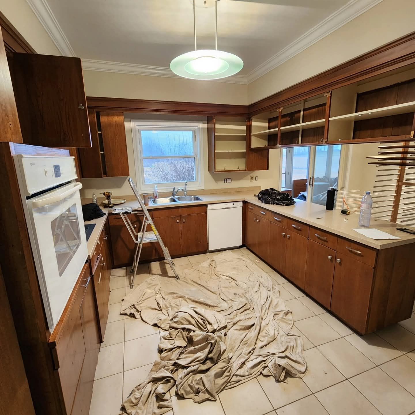 Kitchen undergoing renovation, with brown cabinets, white appliances, and a drop cloth on the floor.