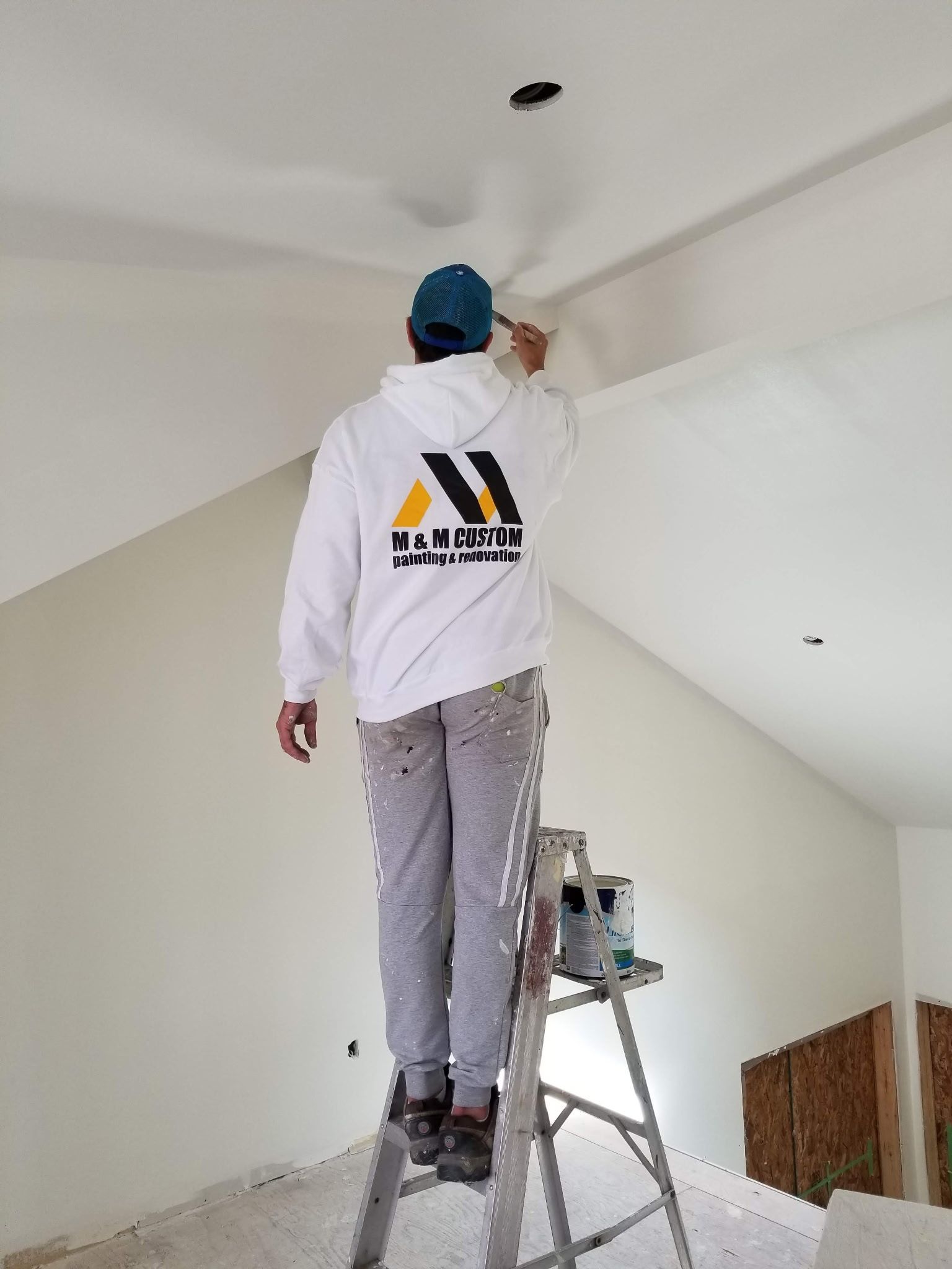 Painter on ladder, painting a white ceiling. He wears a blue hat, white hoodie, gray pants.