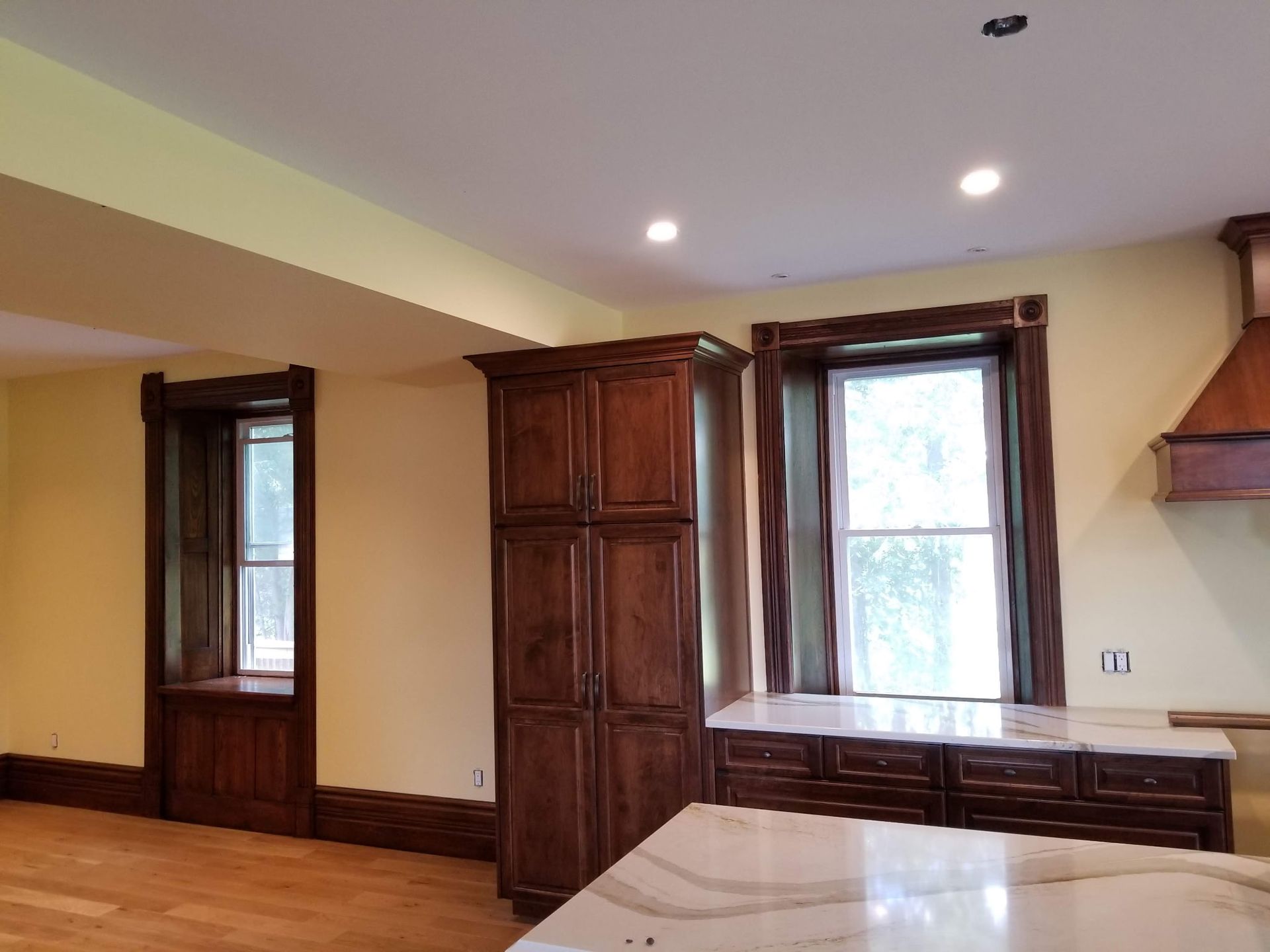 Kitchen with wood cabinets, light countertops, and windows framed with dark wood trim; light yellow walls.