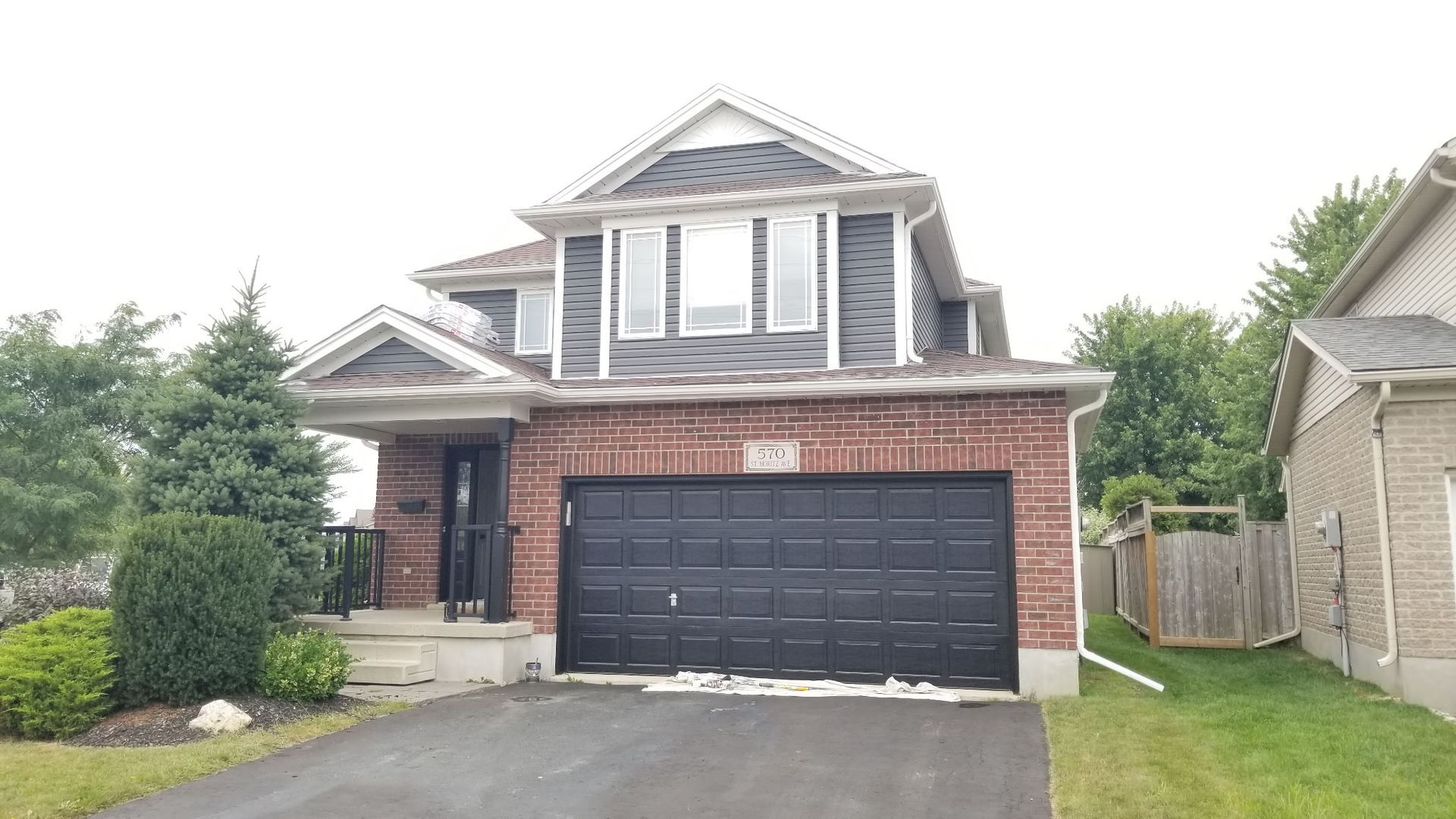 Two-story brick house with black garage door, driveway, and gray siding on the top. Overcast day.