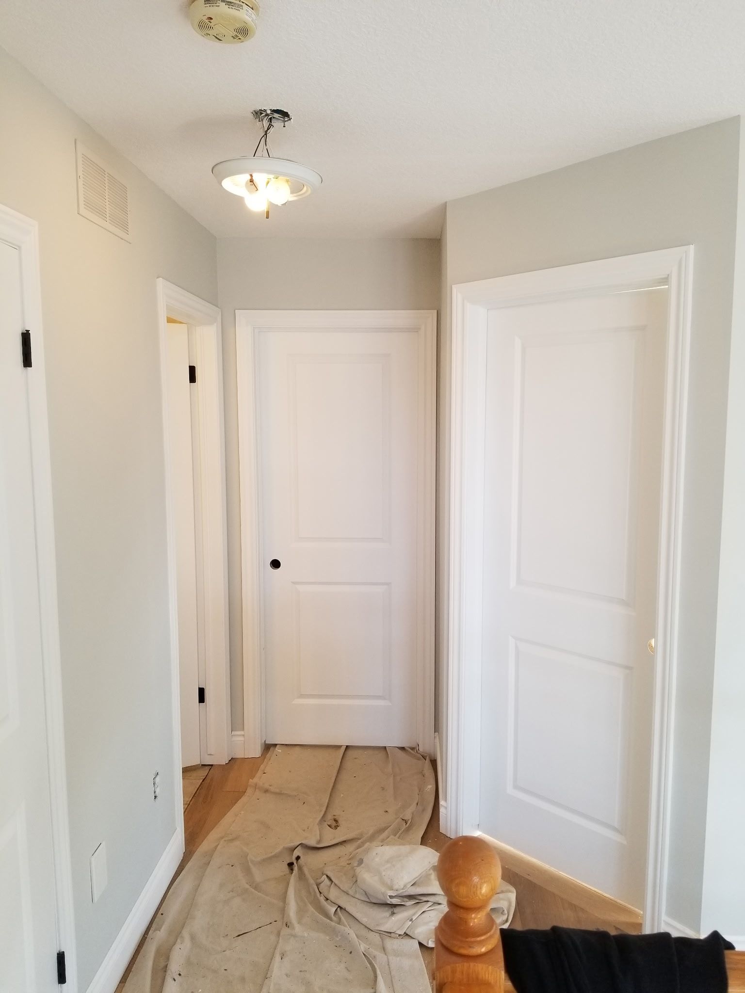 Hallway with three white doors, light gray walls, and a light fixture. Wooden floor with paint tarp.