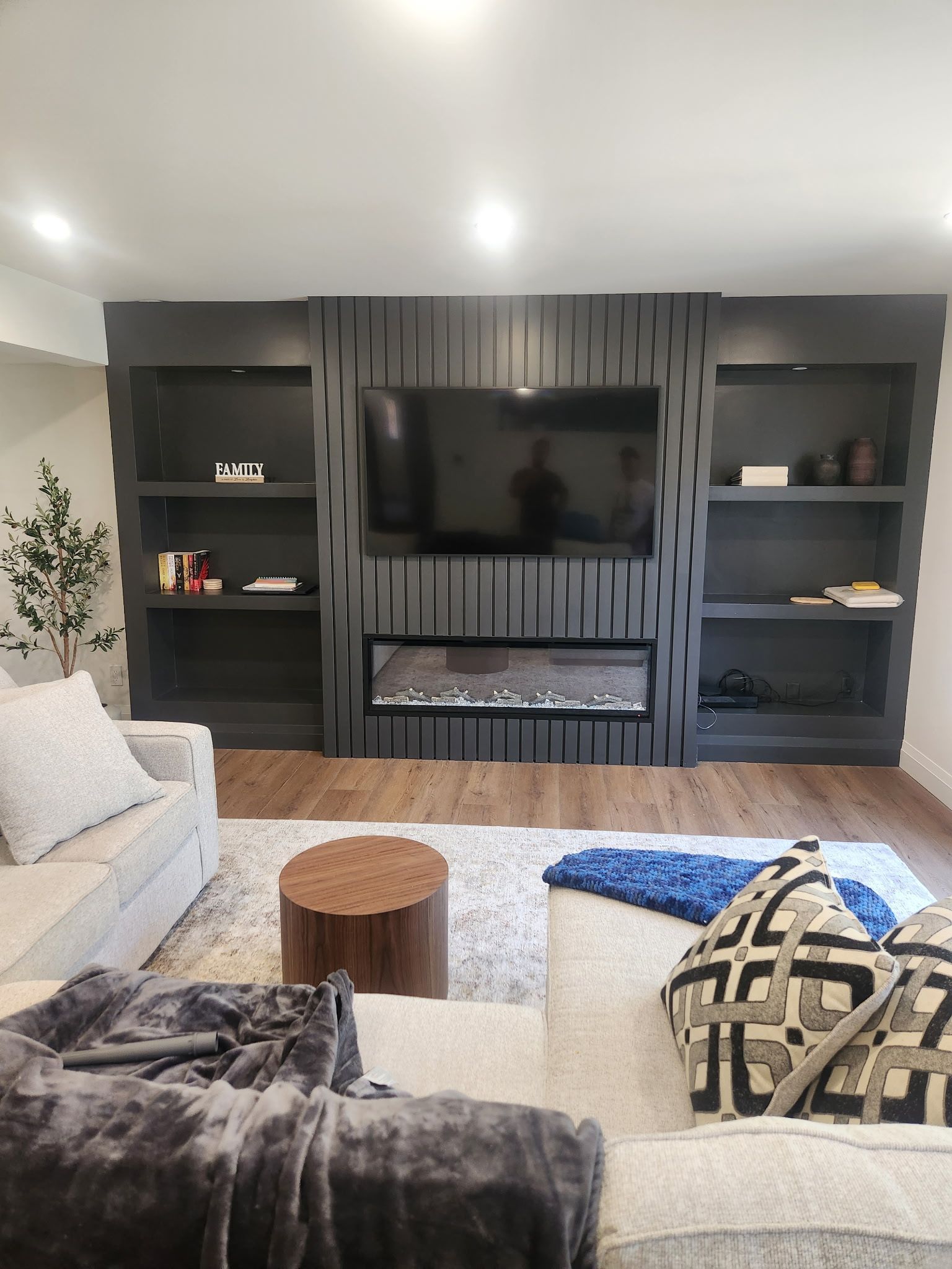Living room with dark gray built-in entertainment center; TV above fireplace, shelving on either side, light sofa, rug.