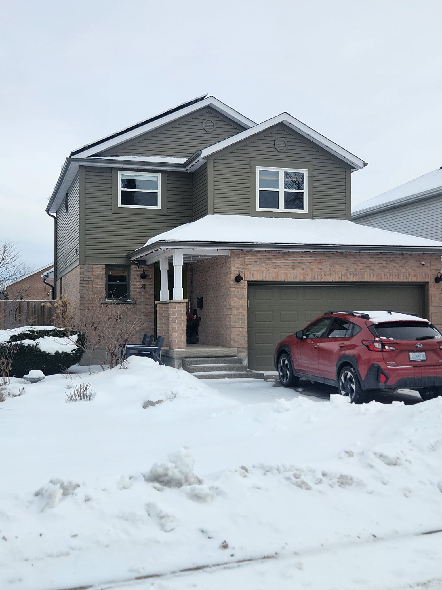 Two-story house with green siding and brick, covered in snow, red car parked in the driveway.