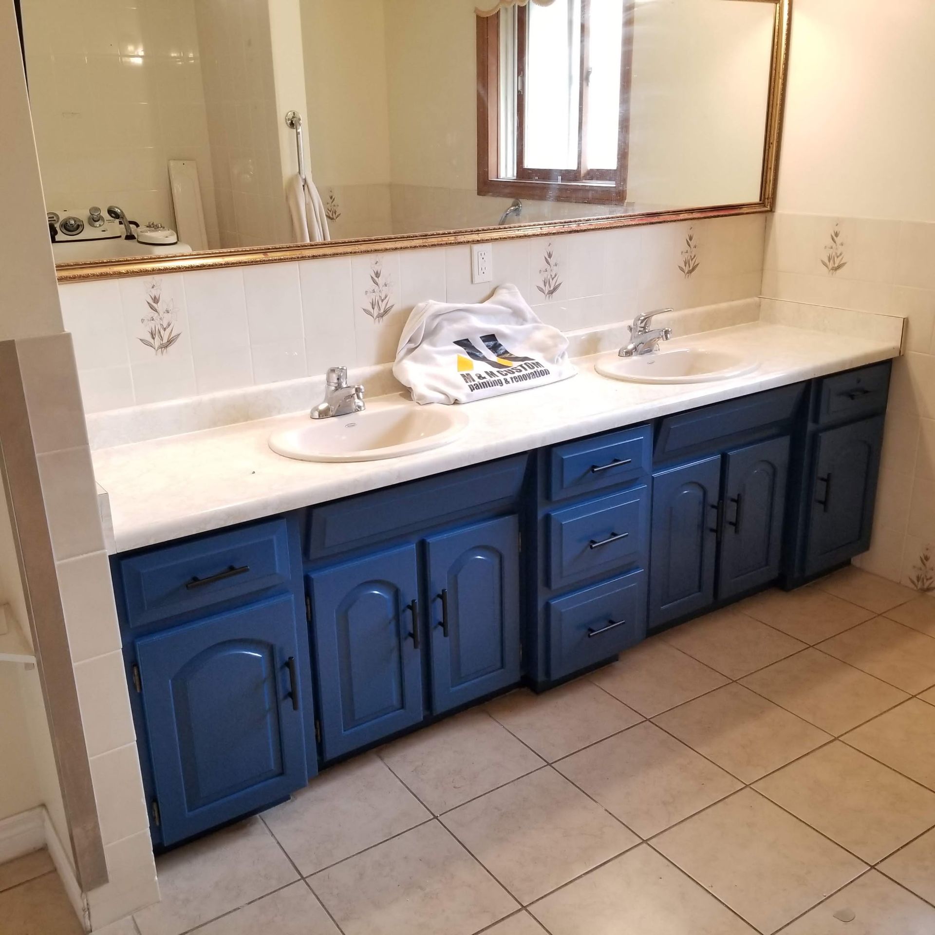 Bathroom with blue cabinets, white countertops, two sinks, and a large mirror.