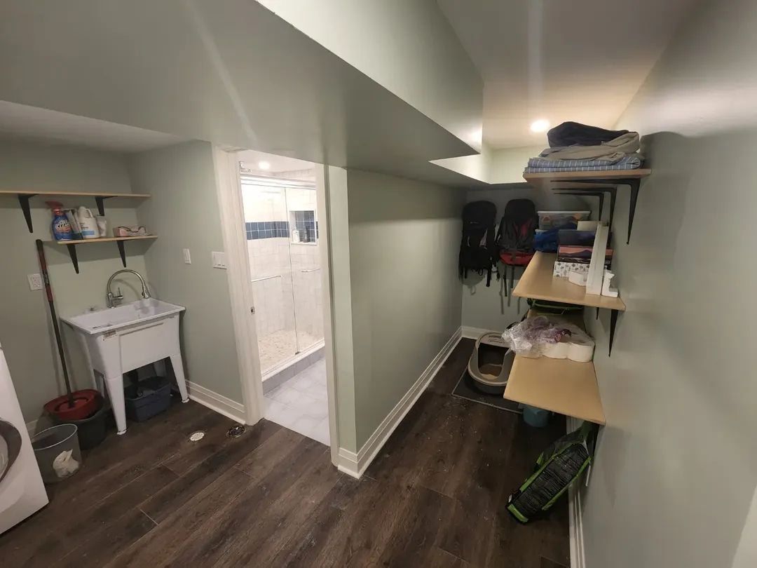 Basement laundry area with sink, shelves, and doorway to a white-walled room. Dark wood-look floor.