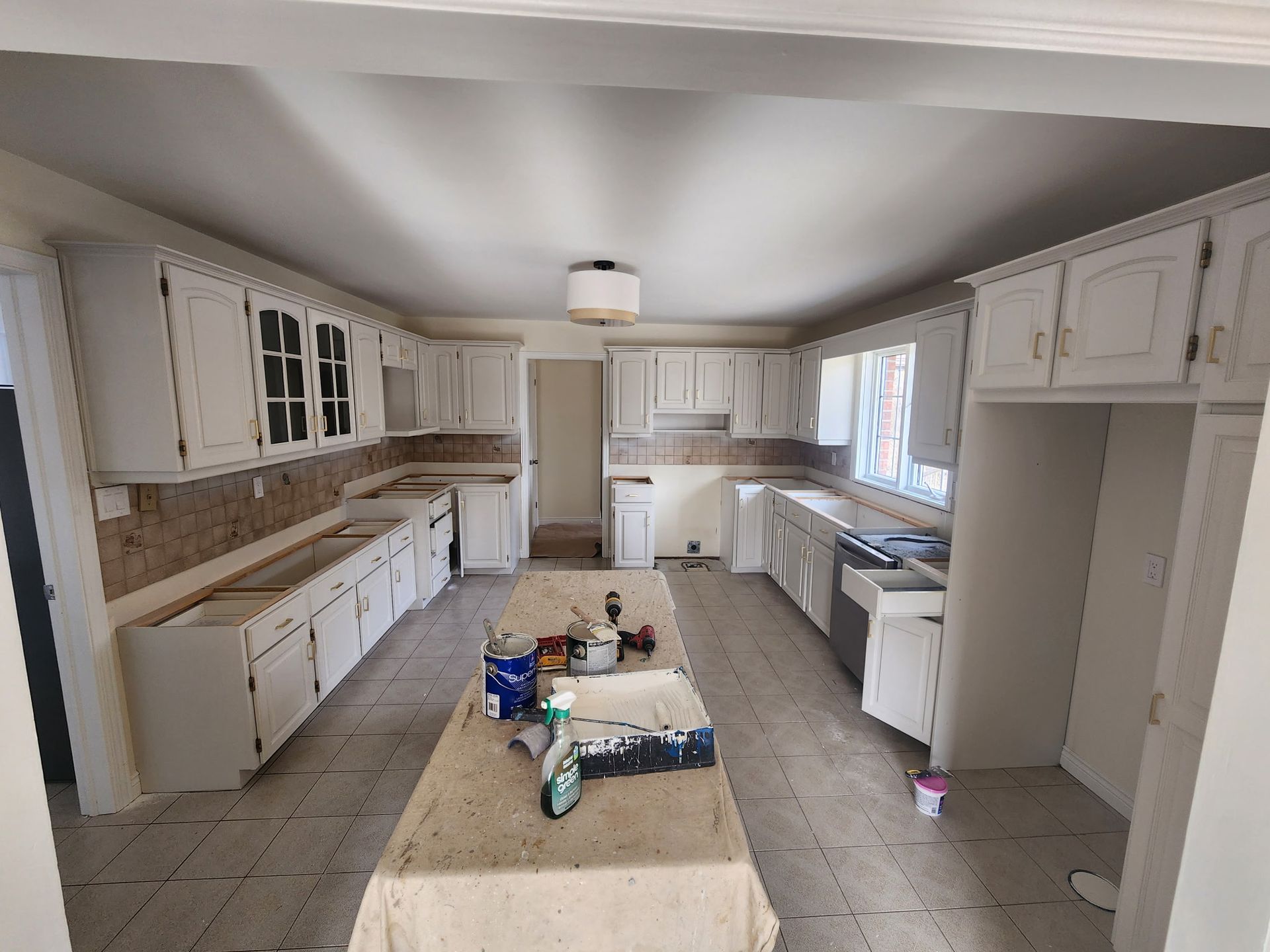 Kitchen undergoing renovation, white cabinets, island with paint supplies, beige backsplash.