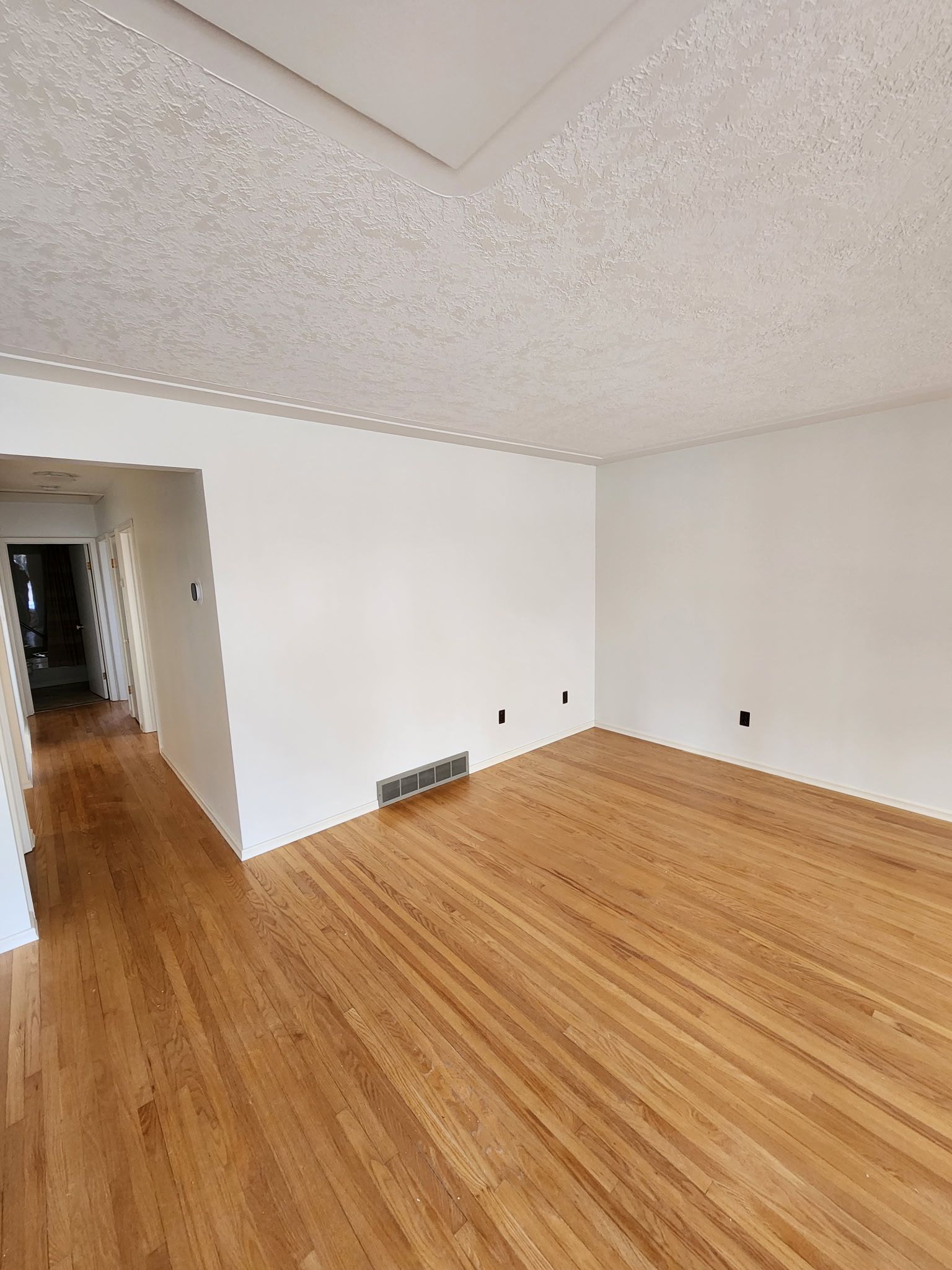 Empty room with hardwood floors, white walls, and popcorn ceiling. Air vent near the floor. Hallway visible.