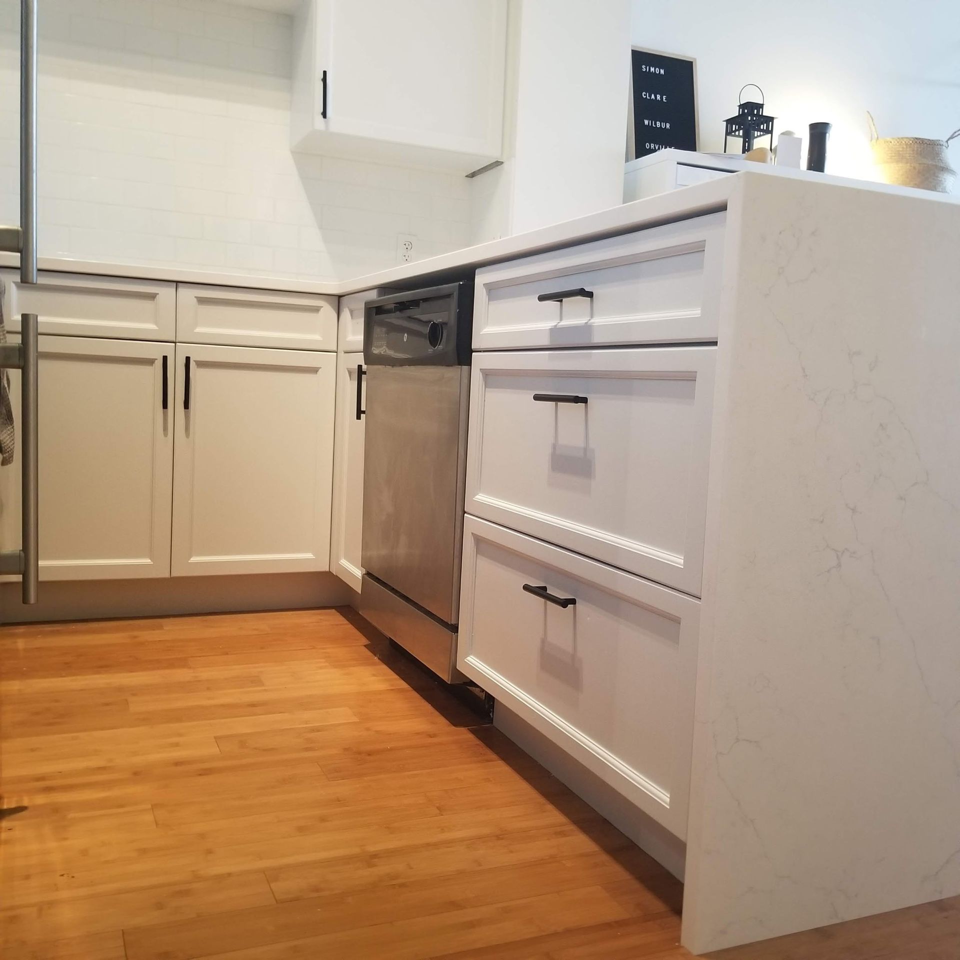 White kitchen cabinets, including an island with drawers, with a dishwasher and light wood flooring.