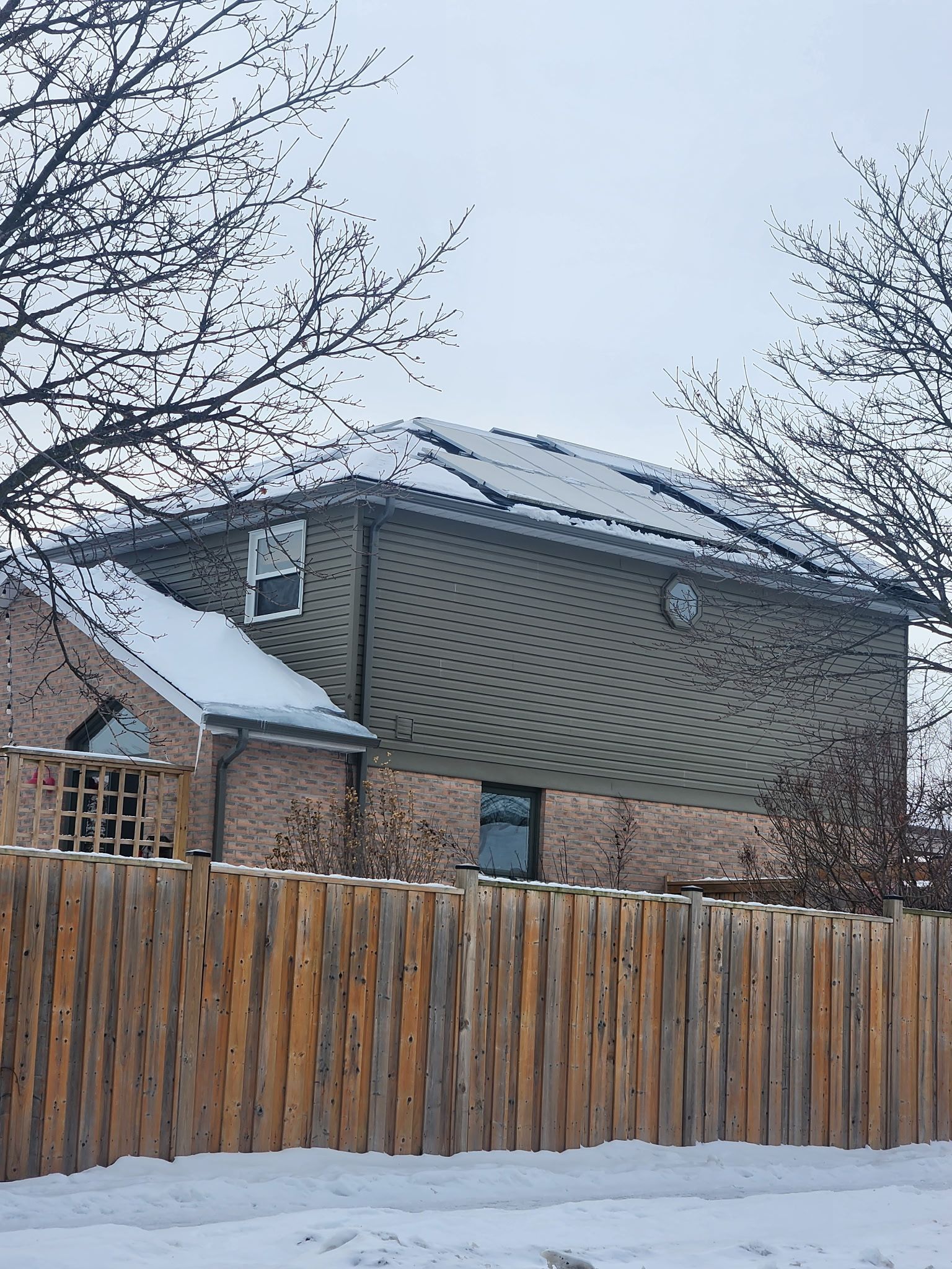 Snowy house with solar panels on roof, behind a wooden fence. Bare trees in the foreground.